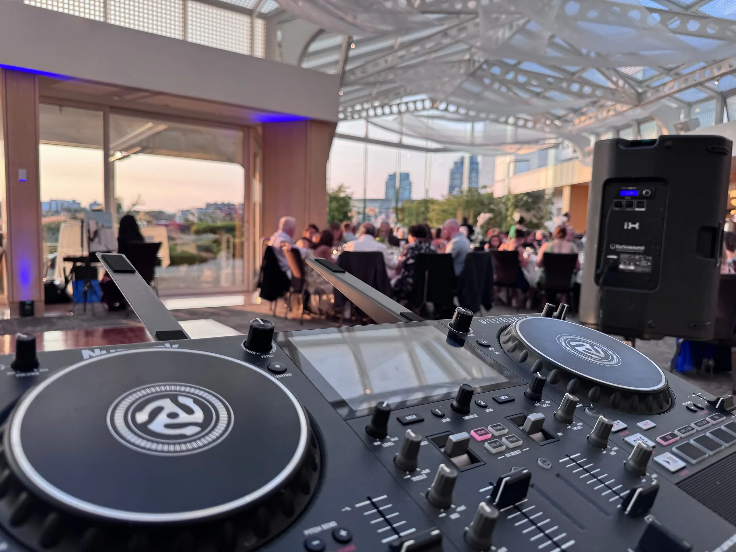 Ryan’s DJ controller and speaker set up at an evening reception, guests dining under a glass roof at sunset