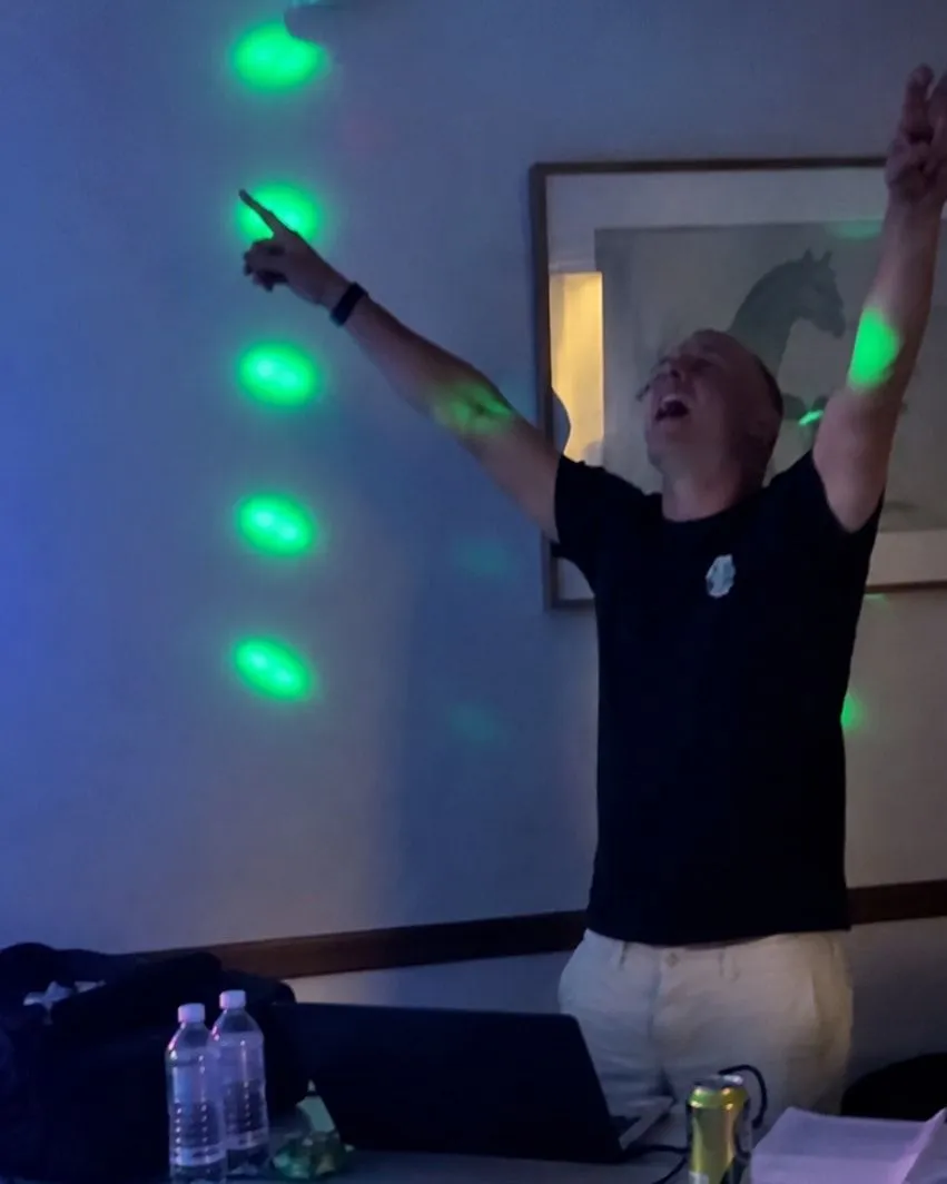Ryan with both arms in the air behind his DJ table, green party lights on the wall