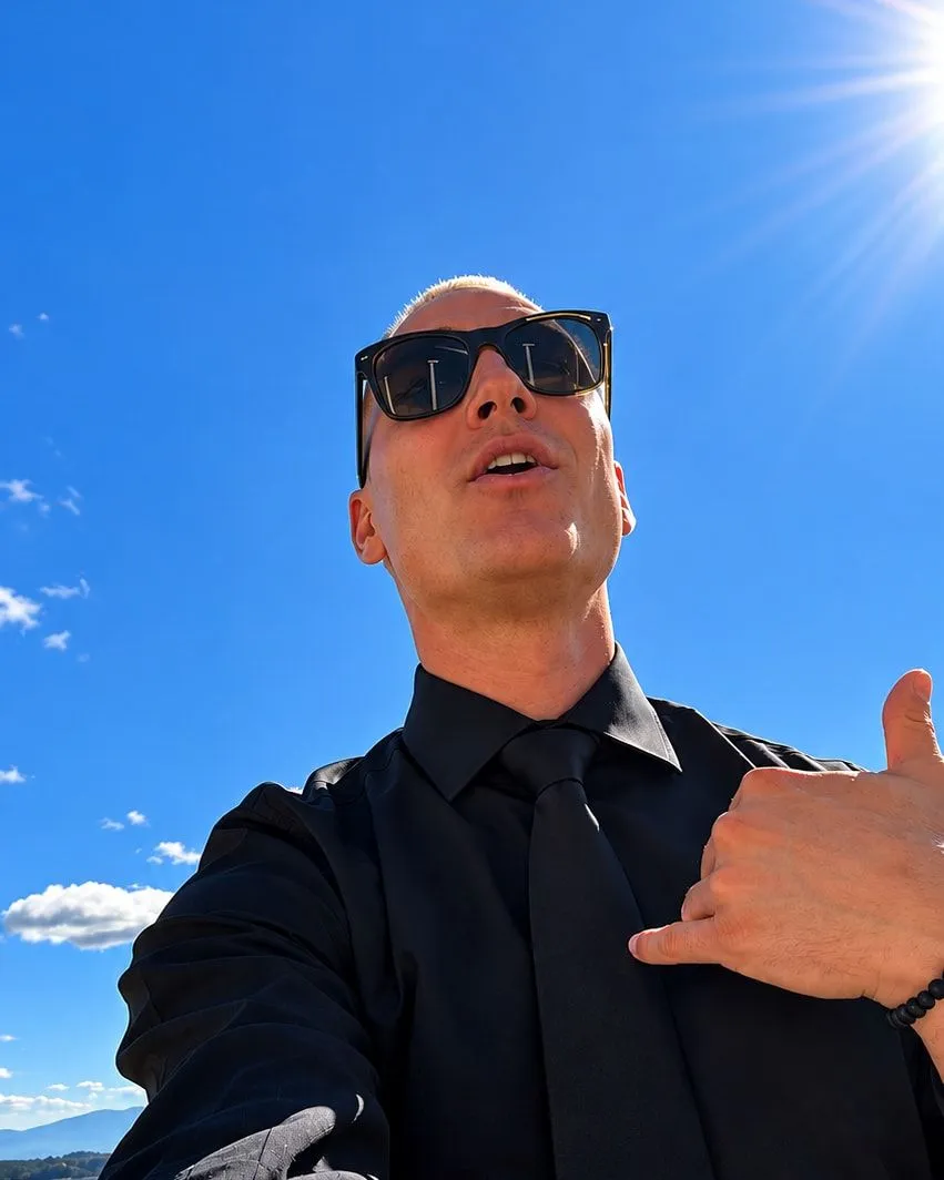 Ryan in sunglasses, a black shirt and tie under a blue sky, giving a shaka sign