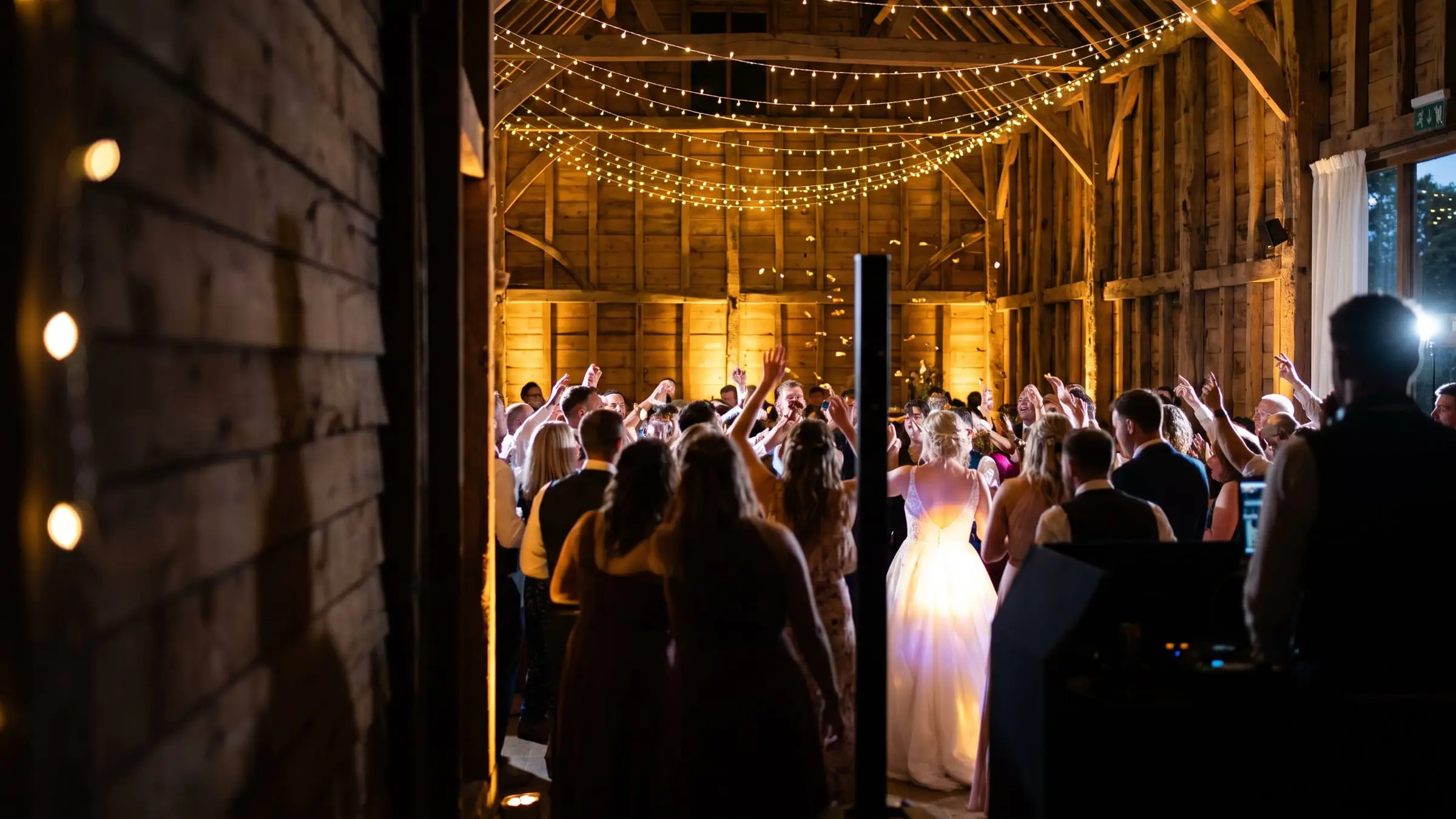 Wedding guests dancing with arms raised under string lights in a timber barn, the bride in white at the centre