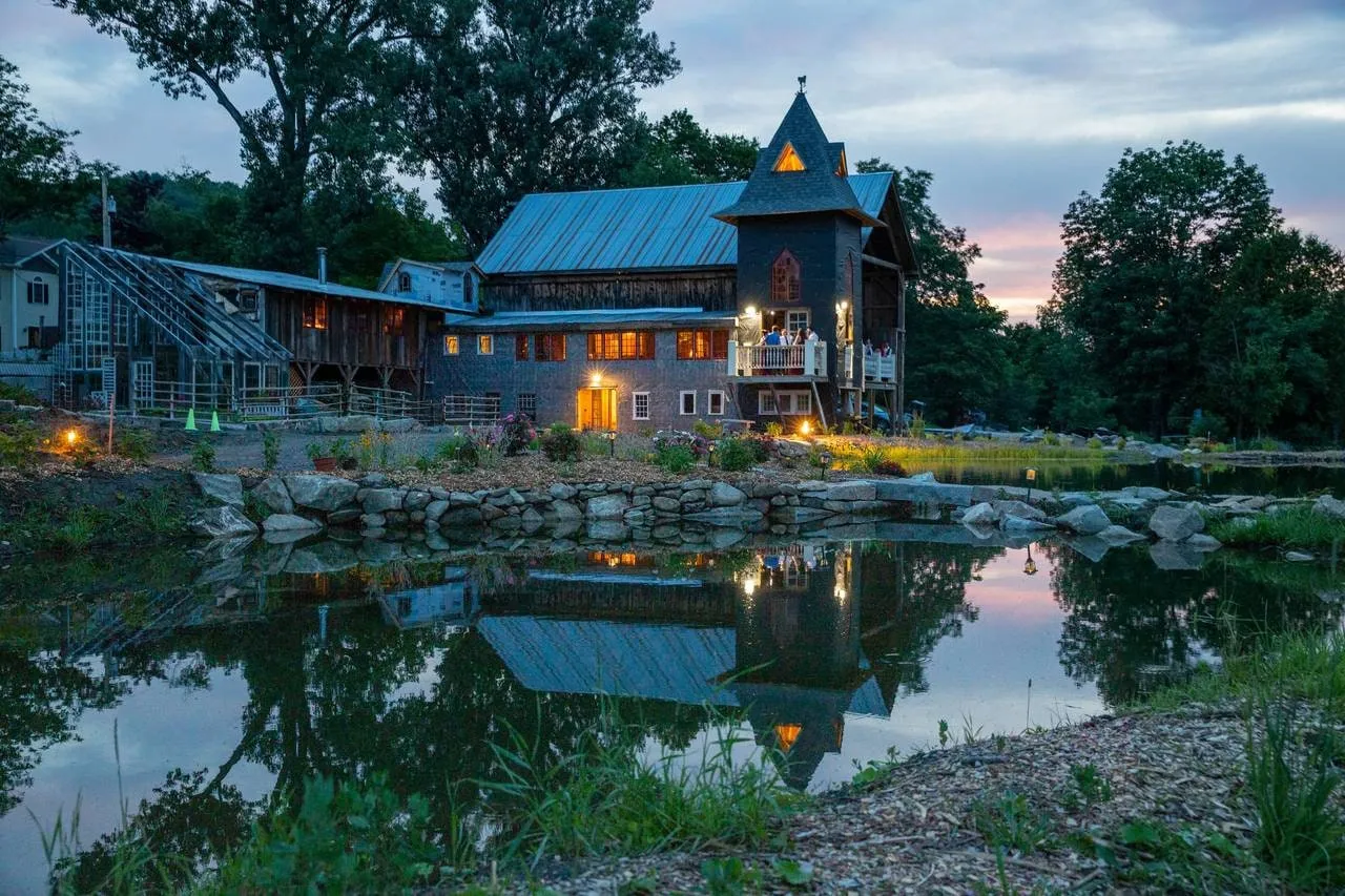 A timber event venue with a steeple tower and lit balcony reflected in a pond at dusk, guests gathered outside