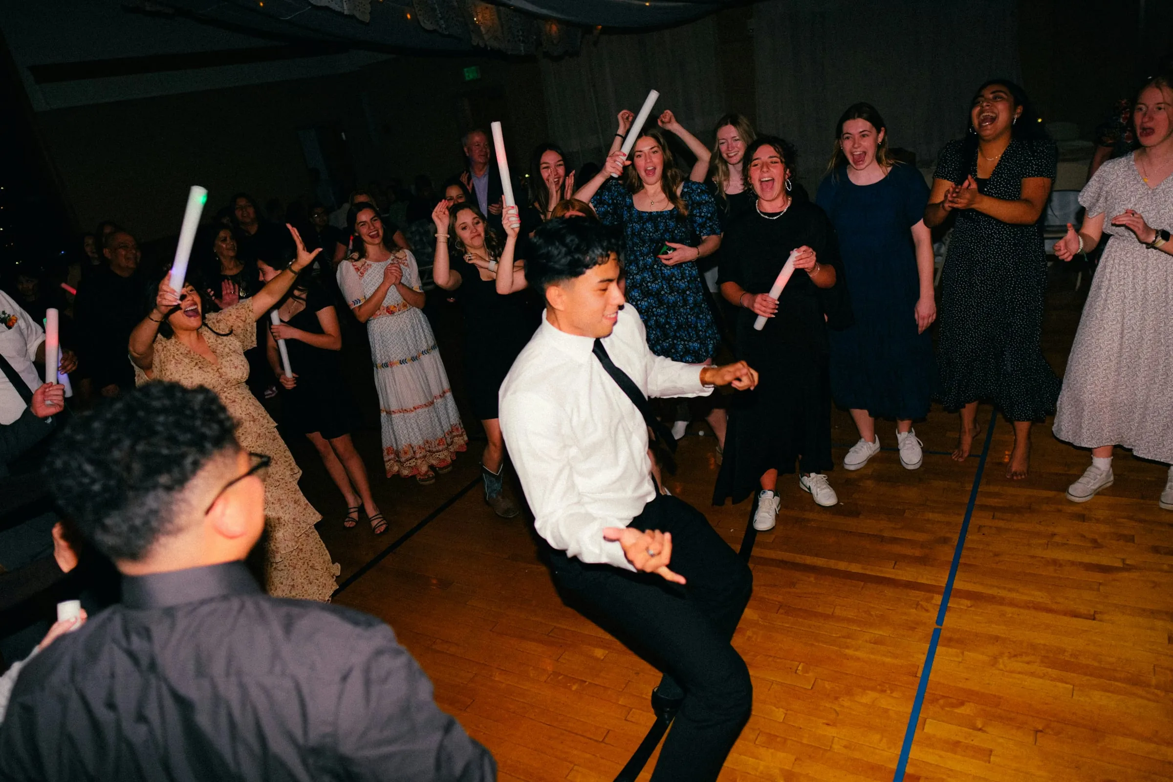 Students cheering with glow sticks as a teen in a white shirt and tie dances on a gym floor