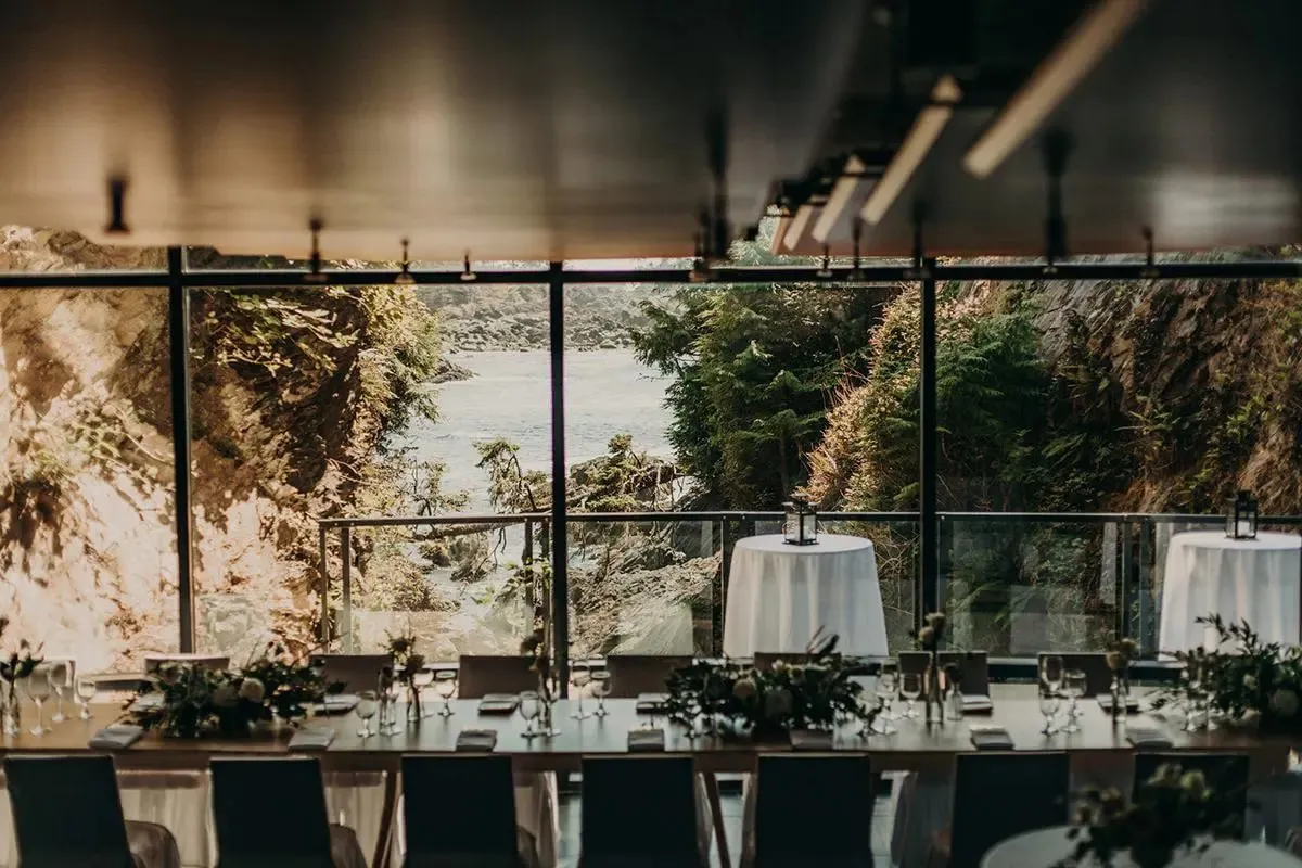 A reception room set with long tables and greenery, floor-to-ceiling windows looking onto a rocky, forested ocean inlet